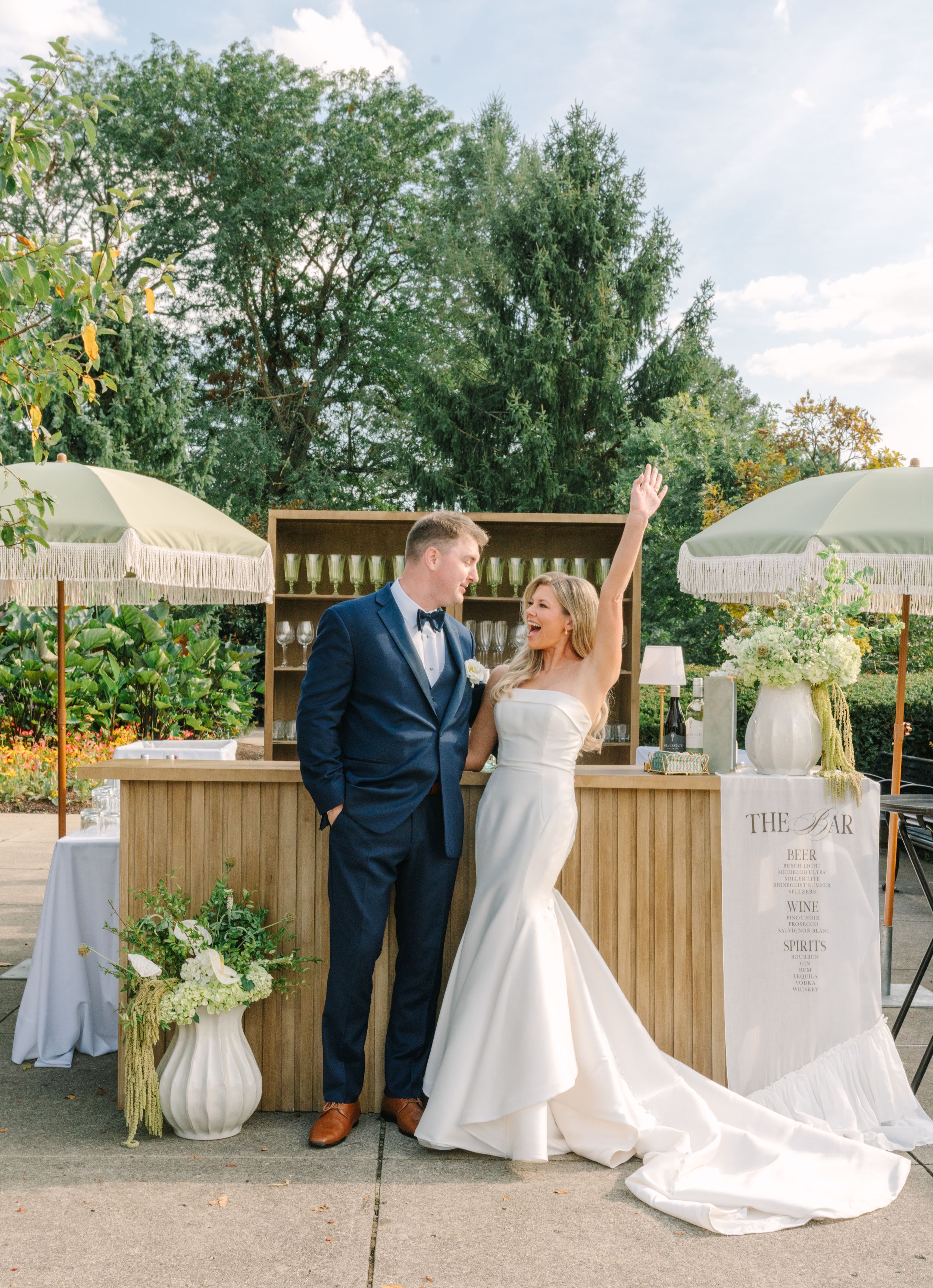 Bride and groom celebrate at outdoor wedding bar with parasols, lush greenery, and elegant white floral arrangements.