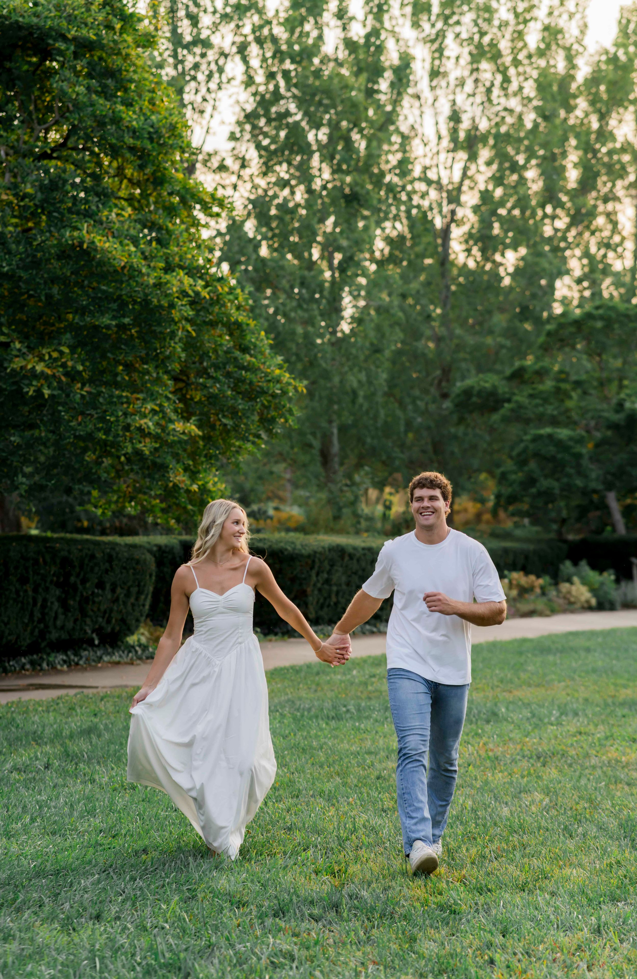 A couple walks playfully across a sunlit lawn with manicured hedges in the background.
