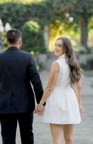 A couple holding hands outdoors in formal attire against a blurred natural background.
