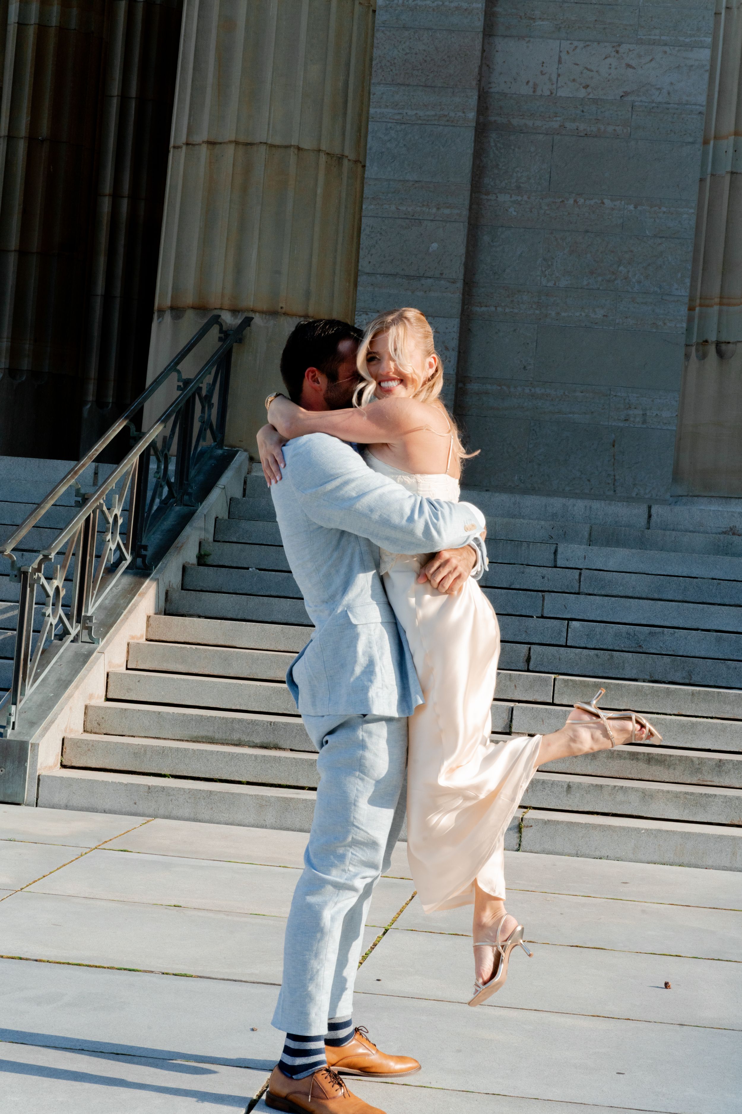 Groom lifting bride in white dress on stone steps outside a grand building on their wedding day.