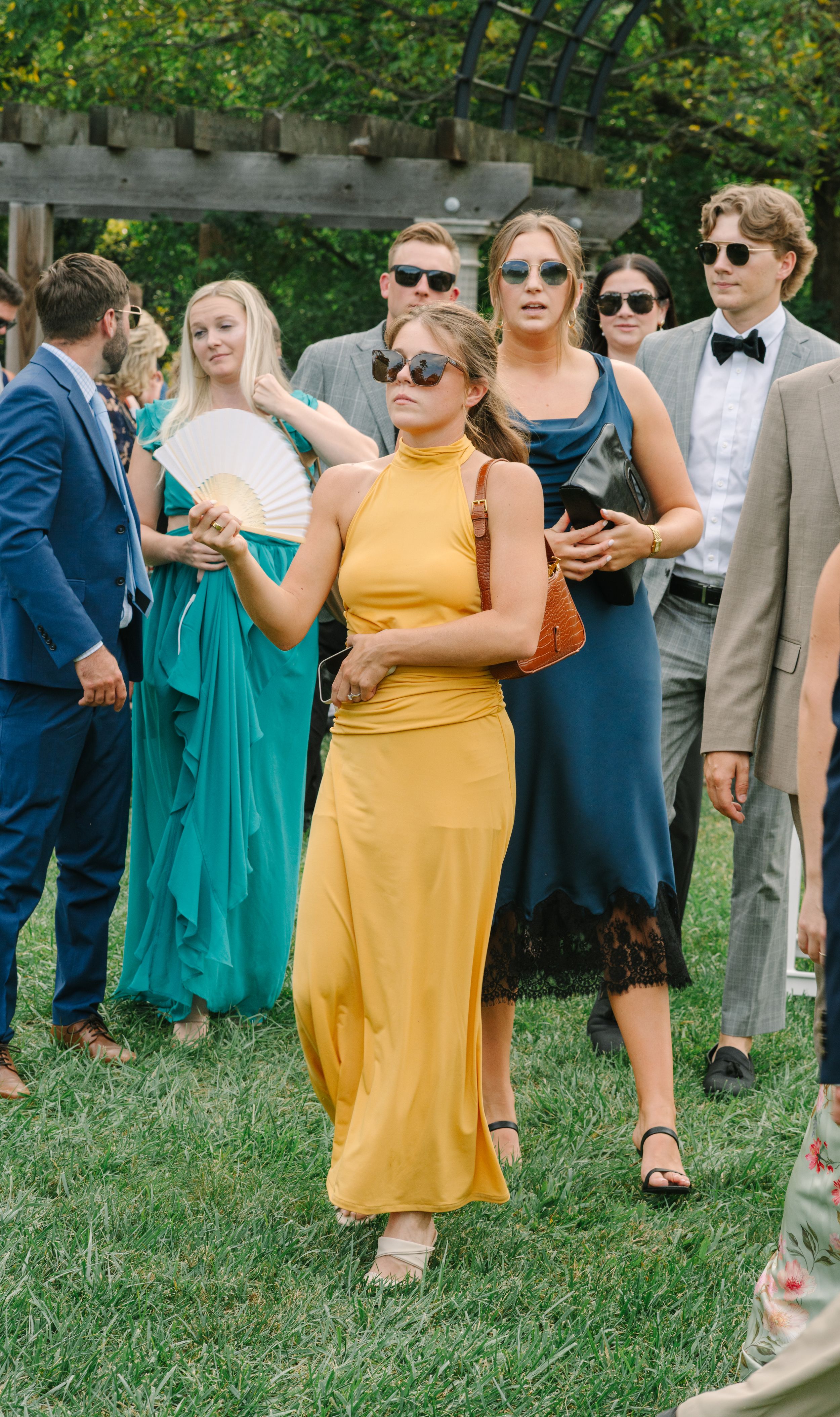 Guests in colorful formal attire gather outdoors at a summer wedding, woman in yellow maxi dress stands out.