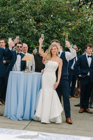 Bride in white strapless gown and groom in navy suit raise champagne glasses with wedding guests outdoors.