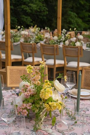 Long dining table set outdoors with pink and yellow floral centerpieces and wooden chairs.