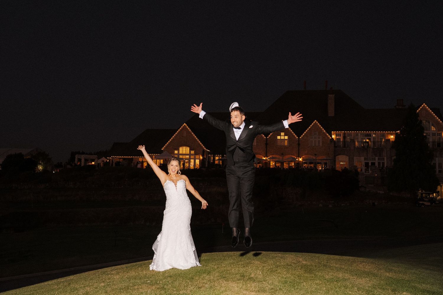 Joyful couple jumps in celebration on a dark golf course with illuminated houses in the background.