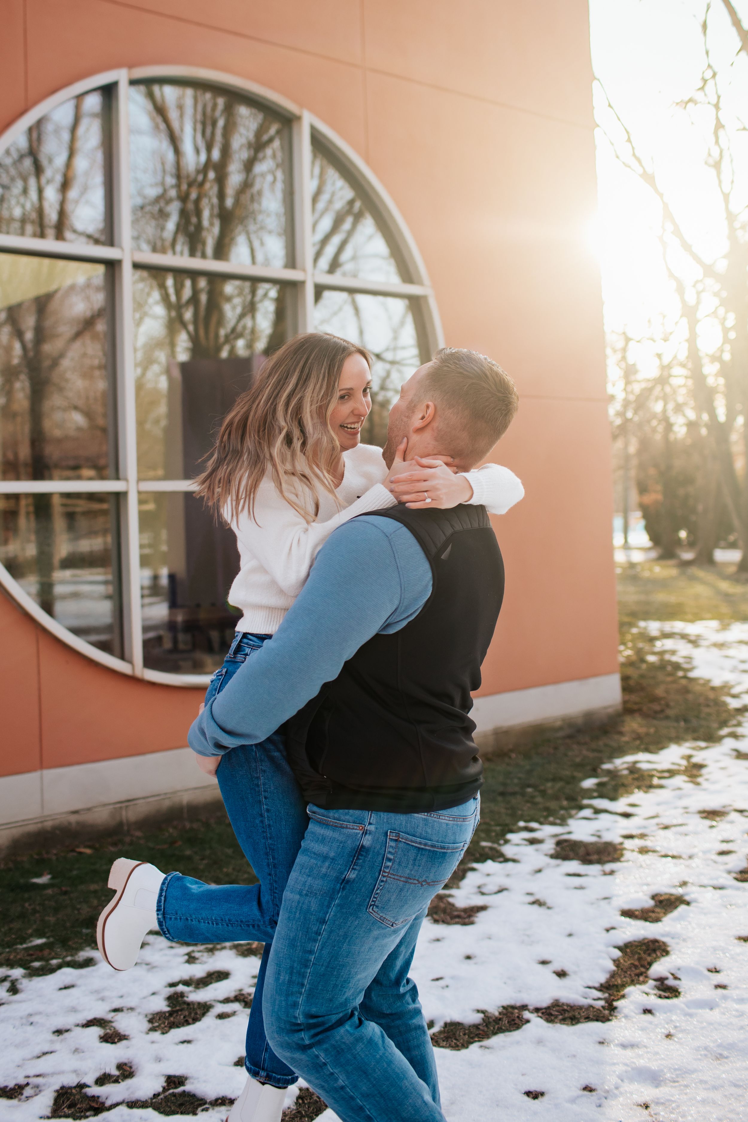 Playful couple embracing outdoors with snow and arched window in background.