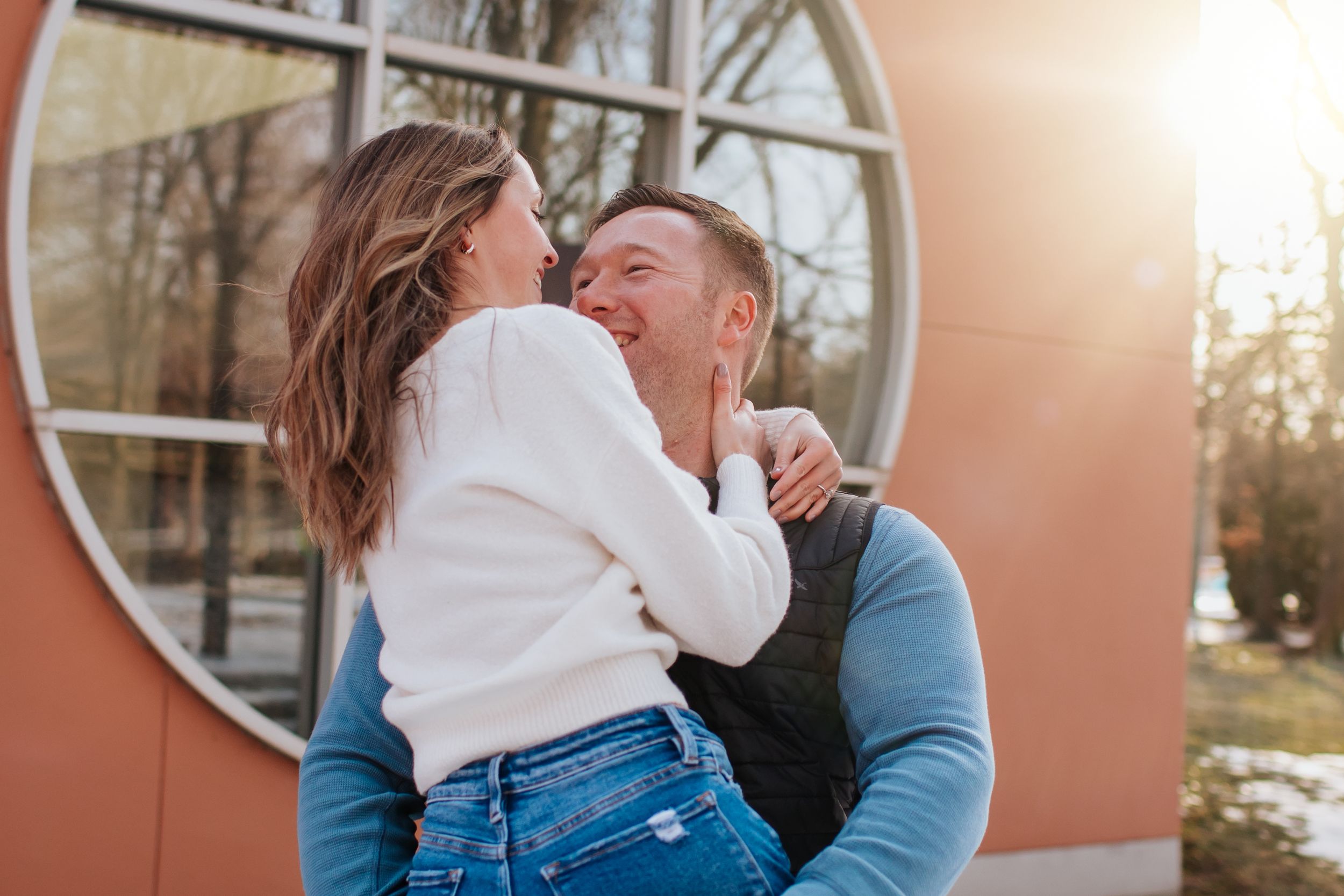 Couple sharing a tender moment outdoors at sunset against an orange wall with circular window.