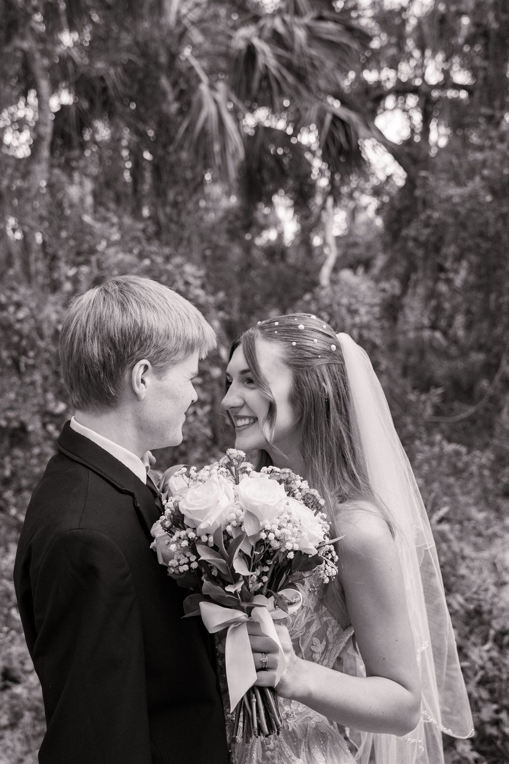 A romantic black and white wedding photo of a couple sharing a joyful moment with a floral bouquet in a garden setting.
