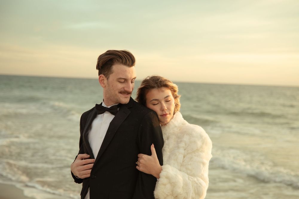 A couple shares a tender embrace on a beach at sunset with waves crashing behind them.