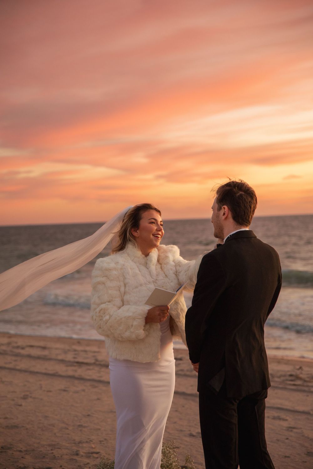 A couple in wedding attire shares a romantic moment on the beach at sunset as the bride's veil flows in the wind.