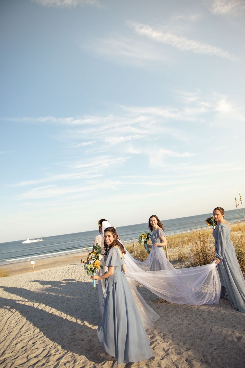 Bridesmaids in flowing blue dresses help adjust a long white veil on a sandy beach at sunset.