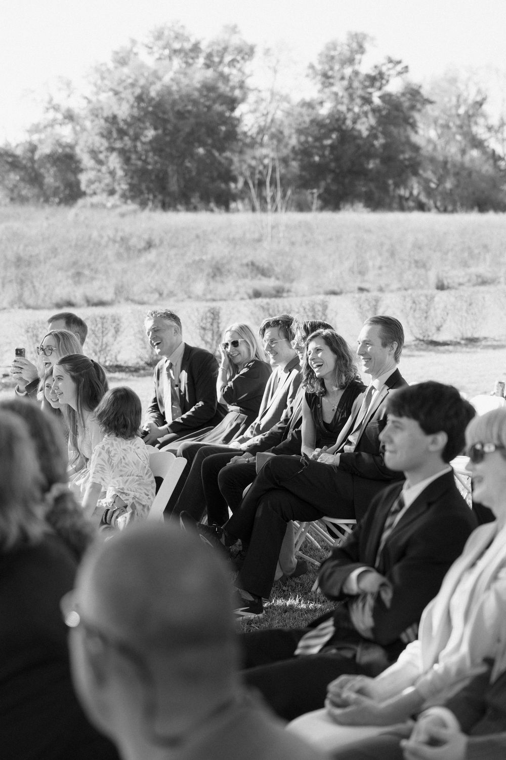 A black and white photo shows a crowd of people sitting in rows at an outdoor event, looking to the side.