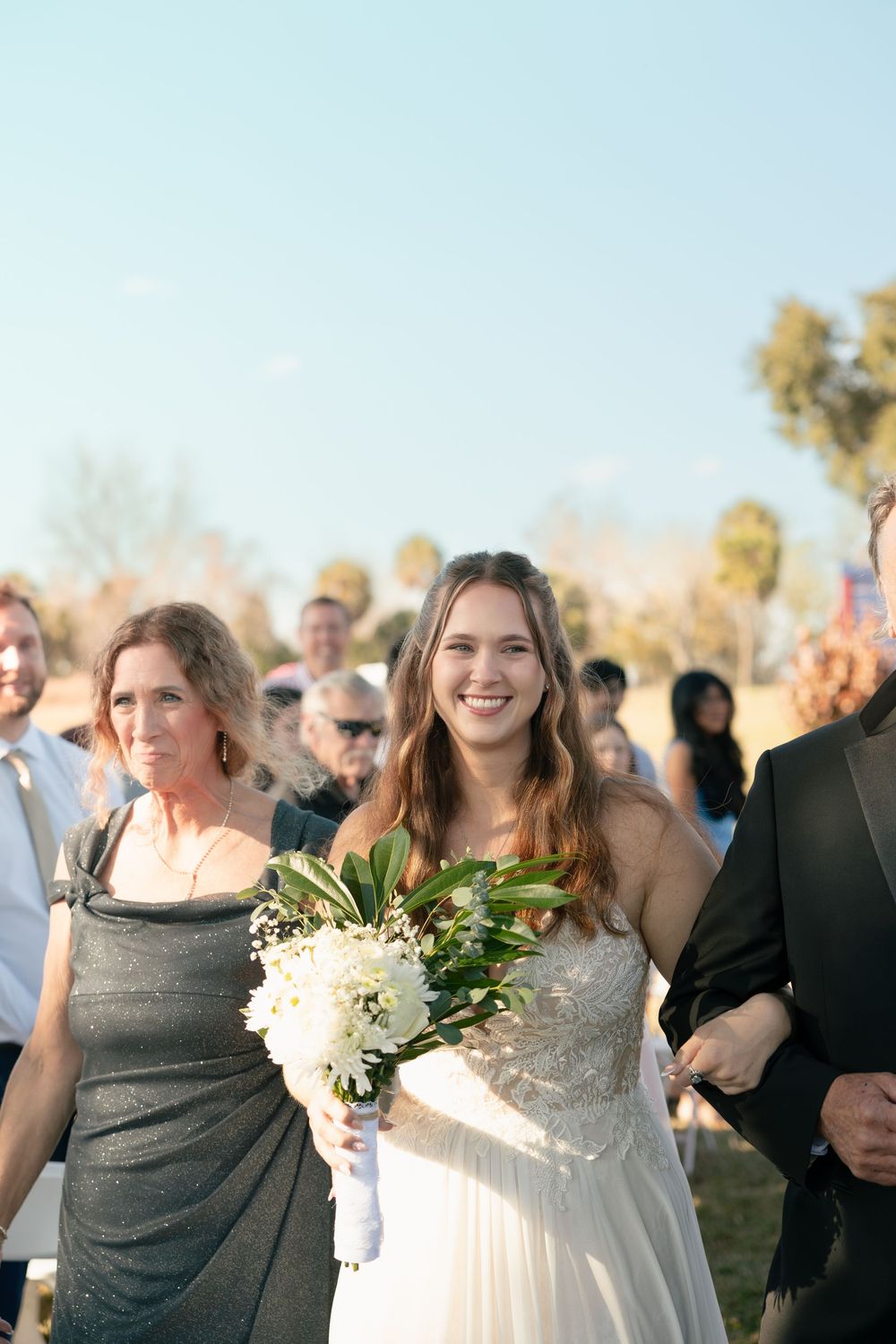 A group of people in formal attire gather outdoors at a wedding celebration on a sunny day.