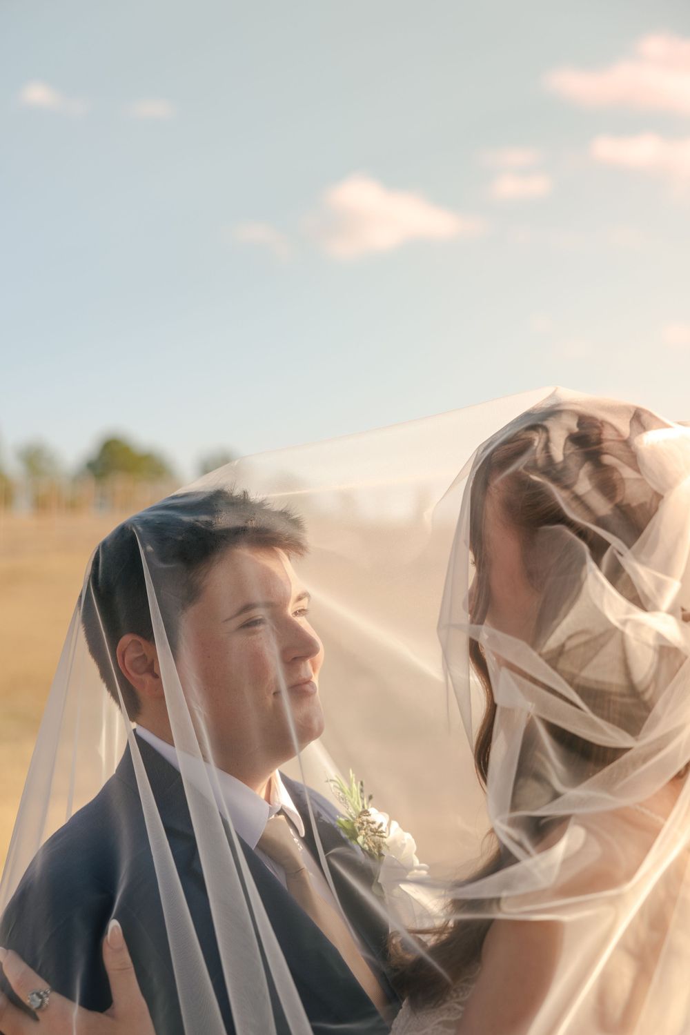 A romantic wedding couple shares a tender moment under a flowing veil against a soft sunset sky.