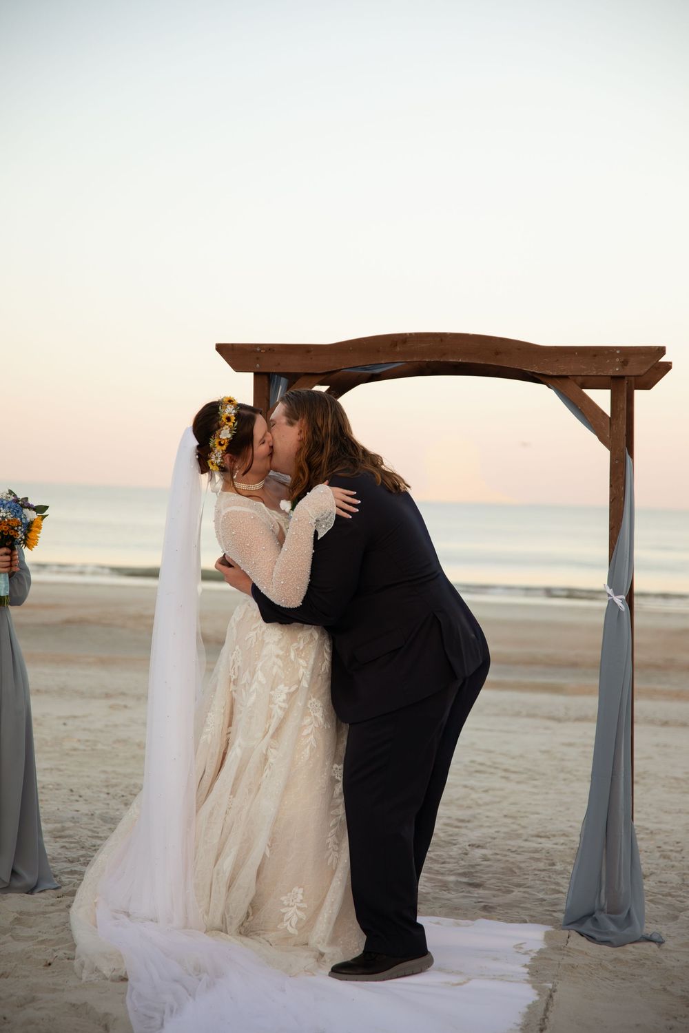 A romantic beach wedding ceremony at sunset with a couple sharing a kiss under a white draped arch on the sand.