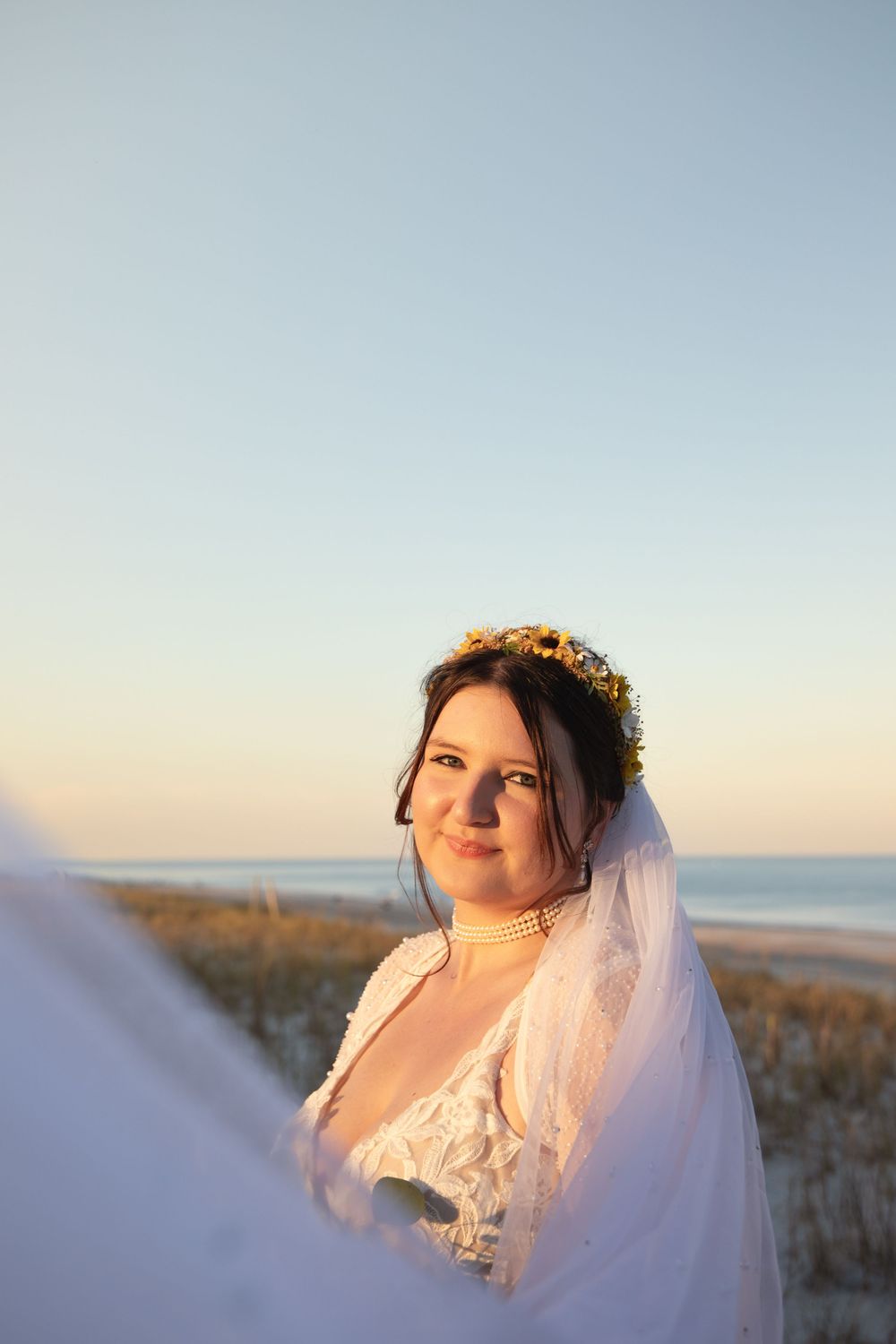 A person in a white wedding dress poses against a soft sunset sky at the beach.