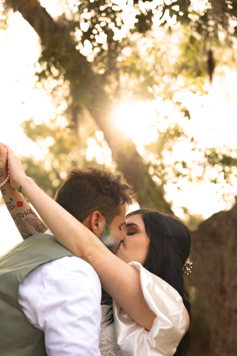 A couple shares a romantic kiss in a sun-dappled garden setting with golden sunlight filtering through trees.