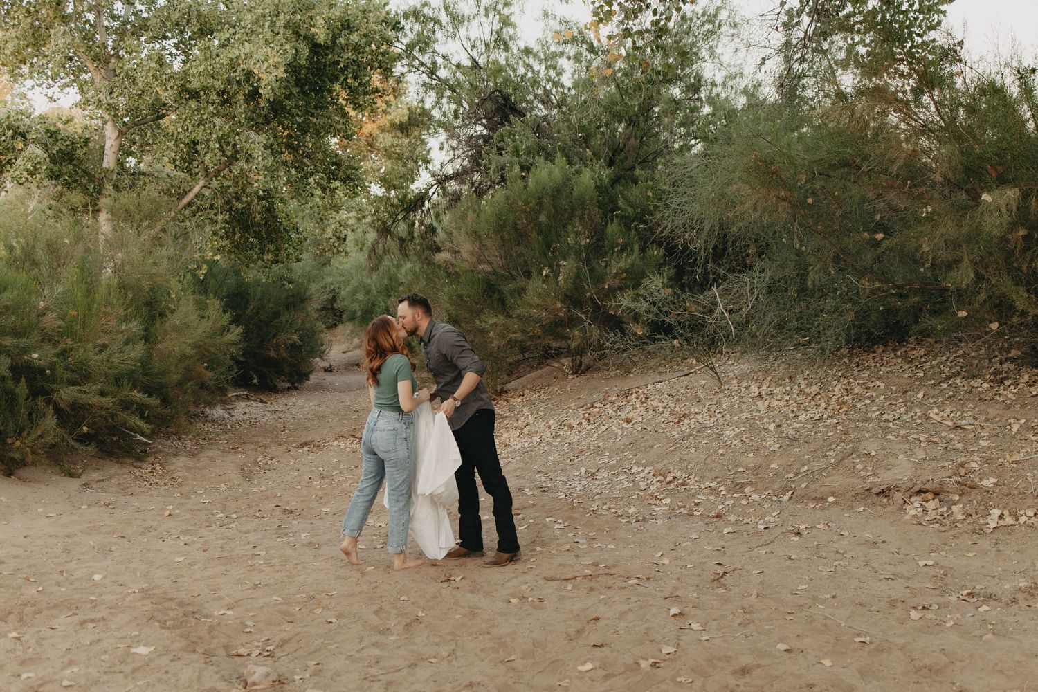 A couple in formal attire stands together on a dirt path surrounded by autumn foliage.