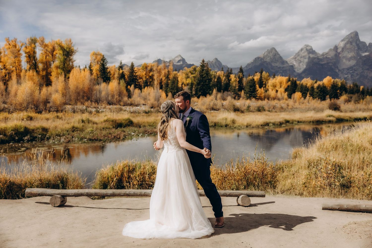 A couple embraces at a scenic overlook with autumn trees and snow-capped Teton mountains in the background.