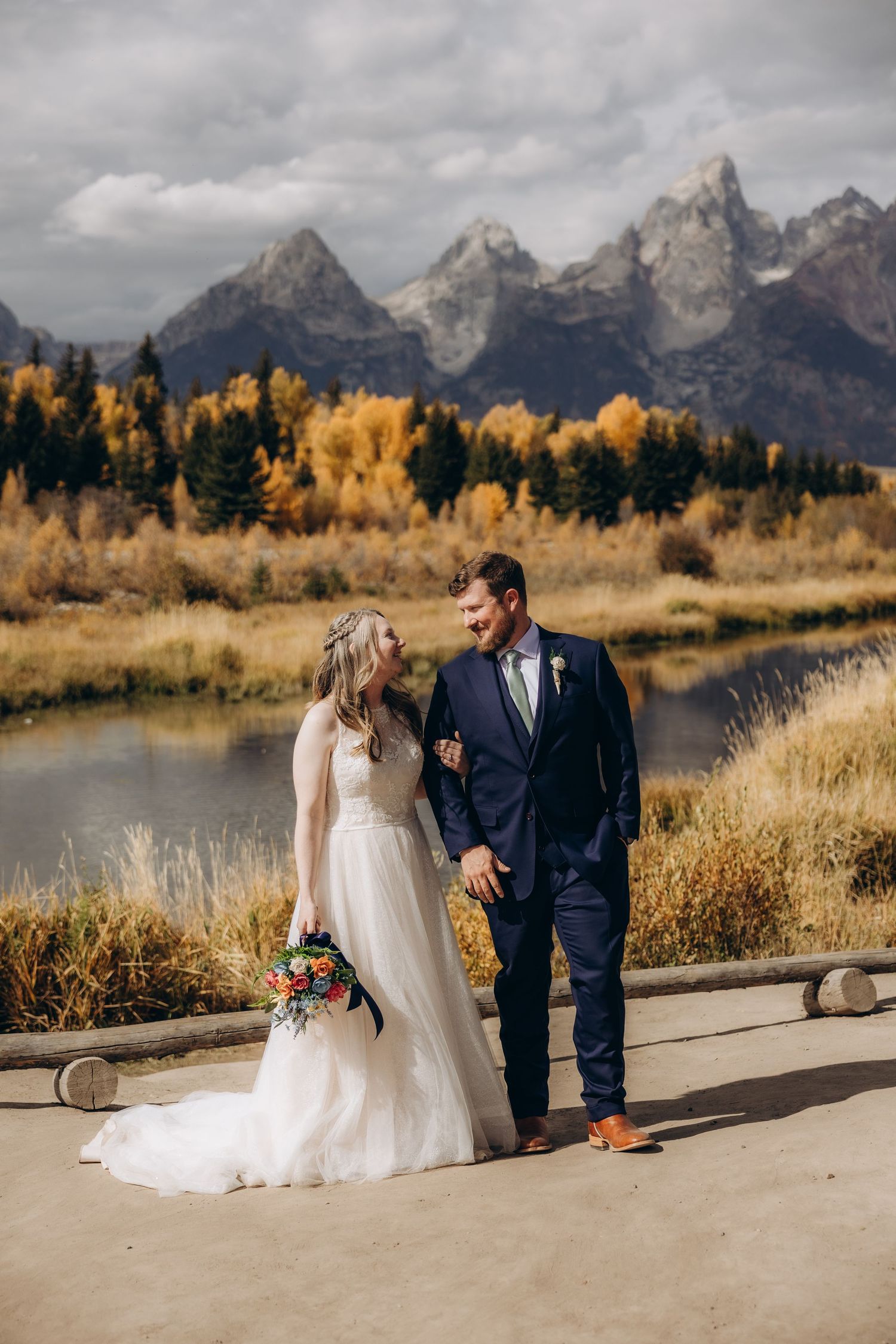 A couple in wedding attire stands together against a backdrop of Grand Teton mountains and autumn foliage in Wyoming.