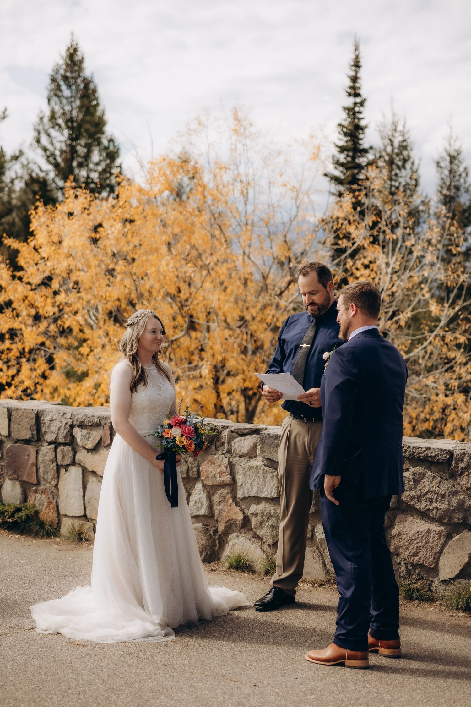A wedding ceremony takes place on a stone path with golden autumn foliage and evergreen trees in the background.