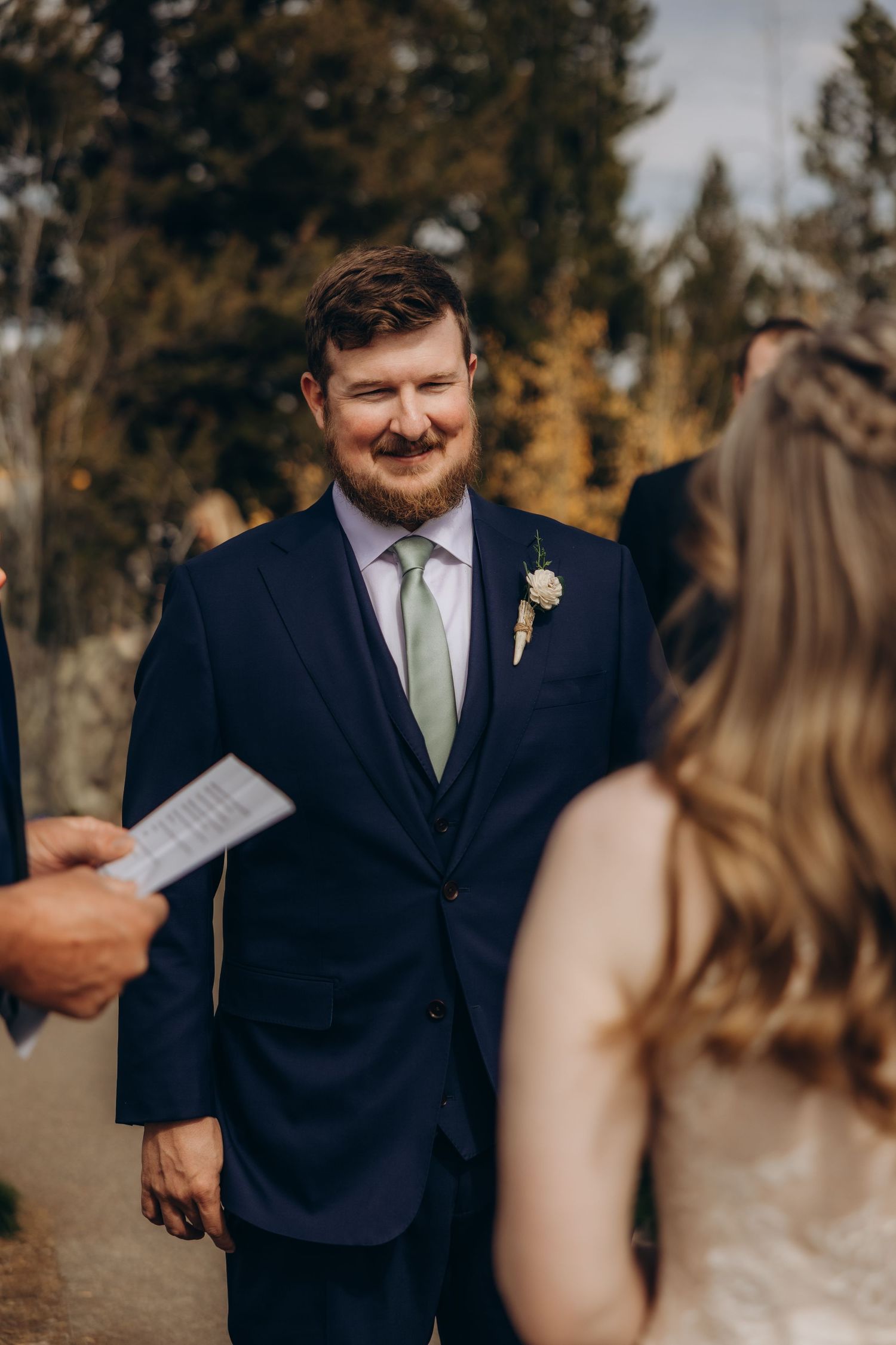 Groom in navy suit with green tie reads vows during outdoor autumn wedding ceremony.