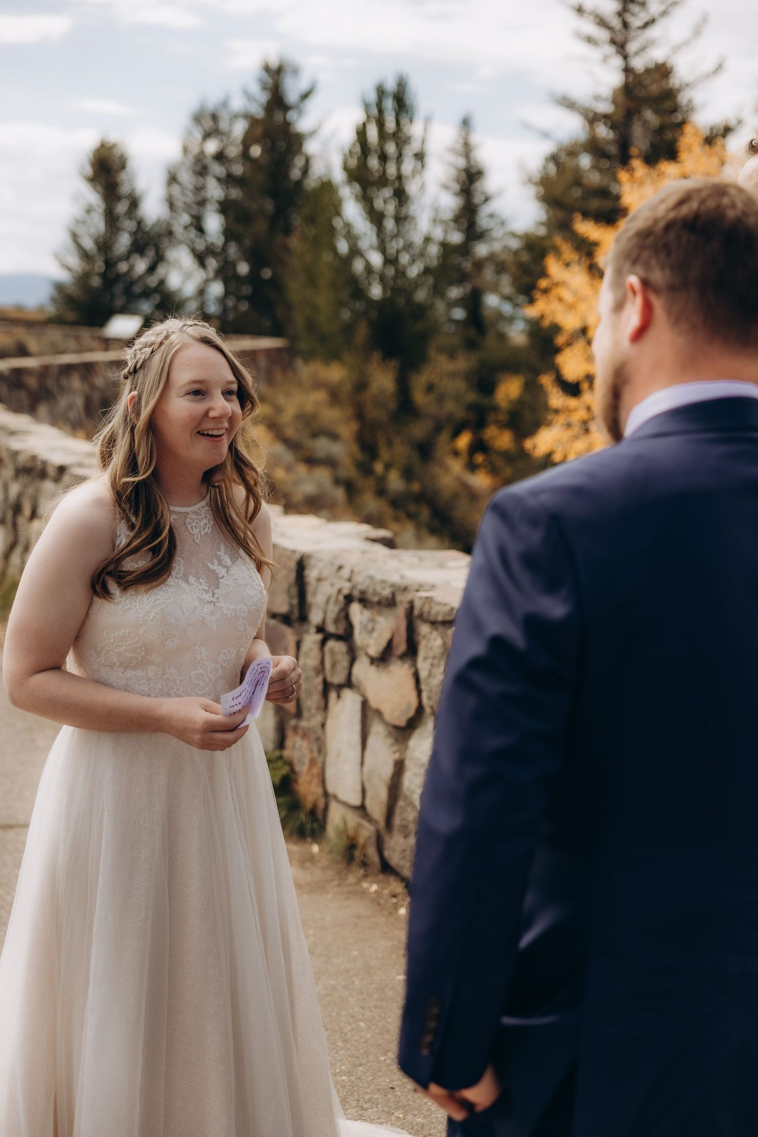 Bride in white lace dress laughs joyfully during outdoor wedding ceremony with fall foliage.