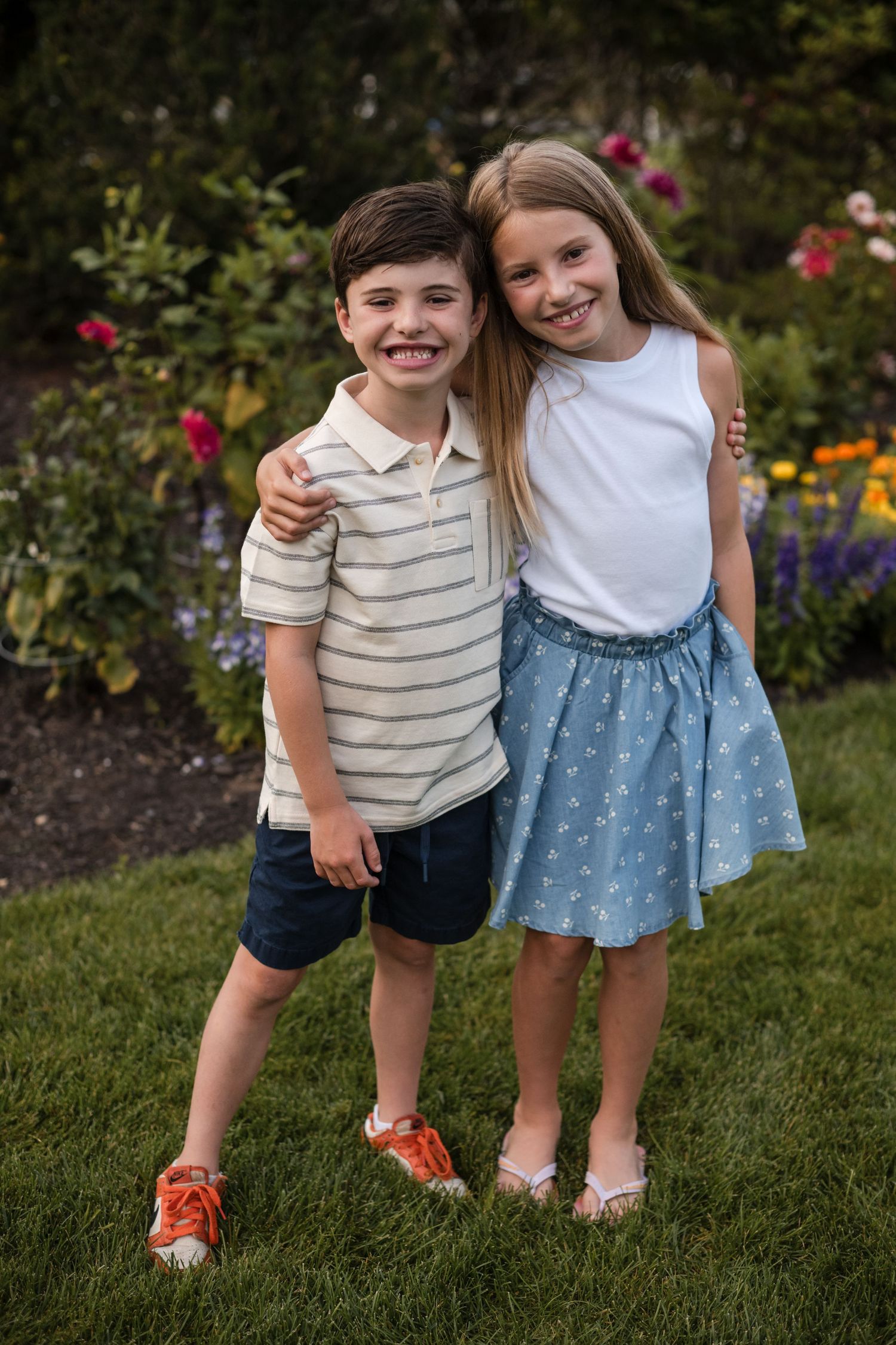 Two children stand together on green grass in a garden with blooming flowers in the background.