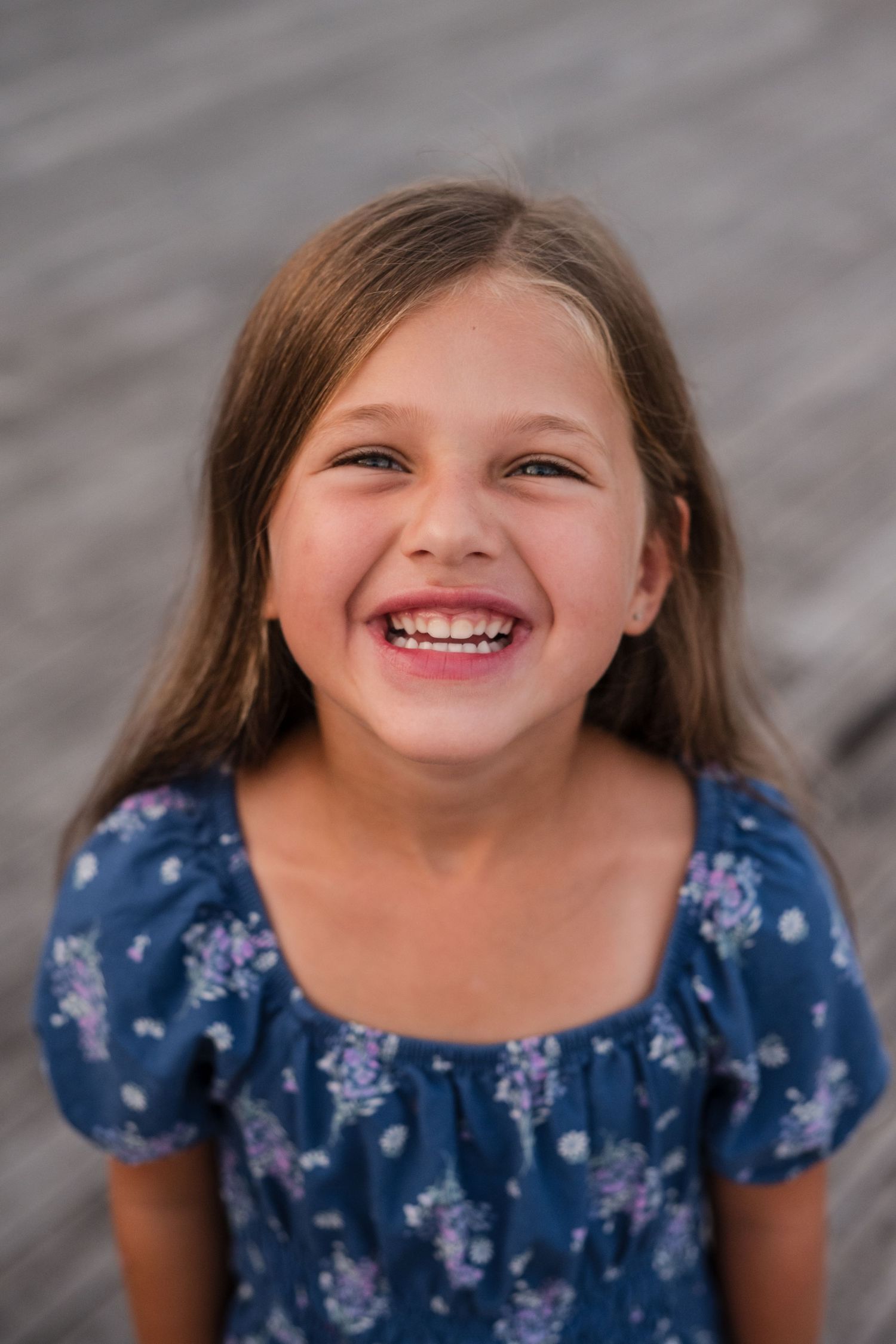 A young child wearing a blue floral dress smiles brightly against a gray background.