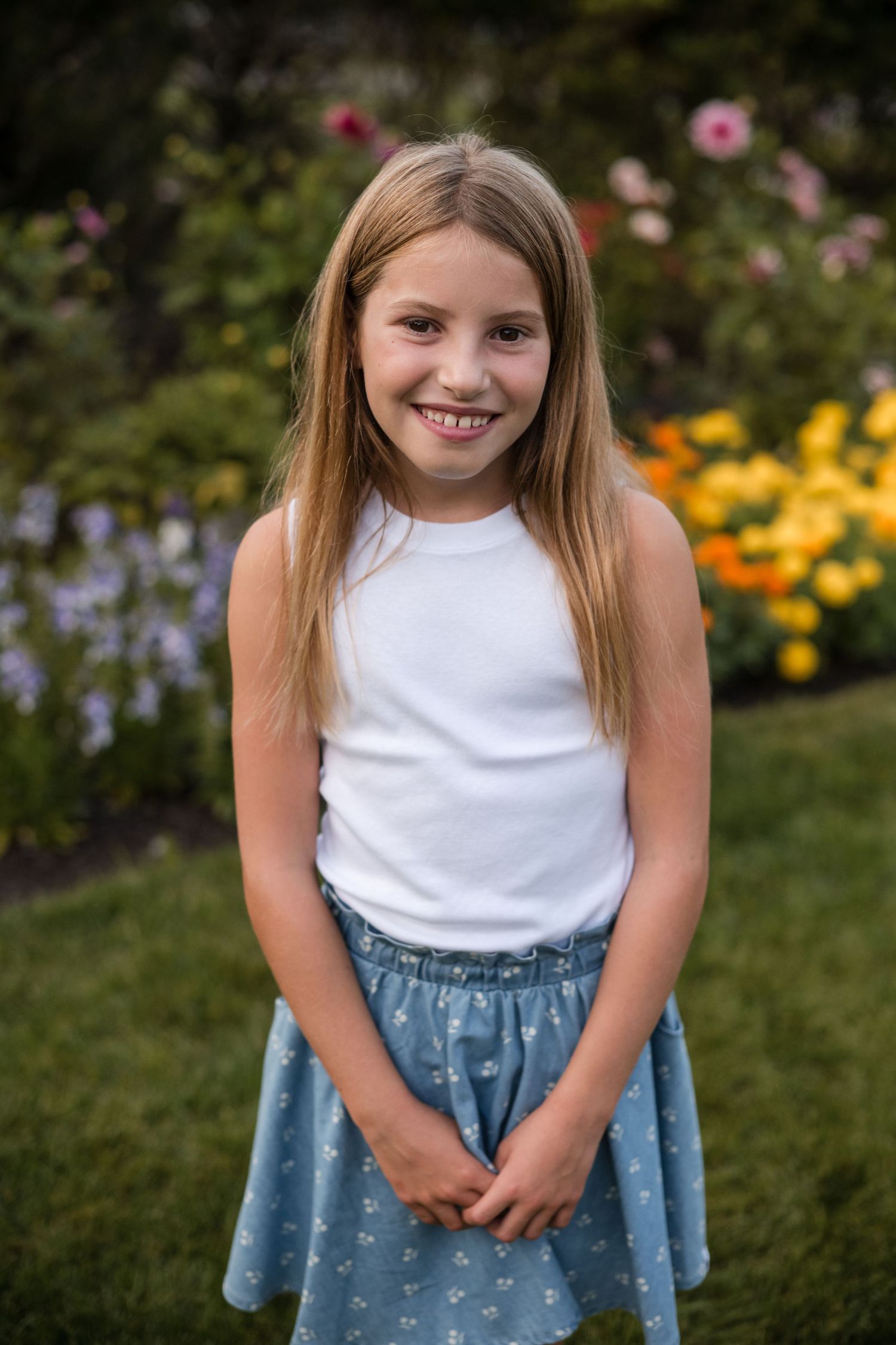 A young girl in a white tank top and floral denim skirt poses cheerfully in a garden with colorful flowers.