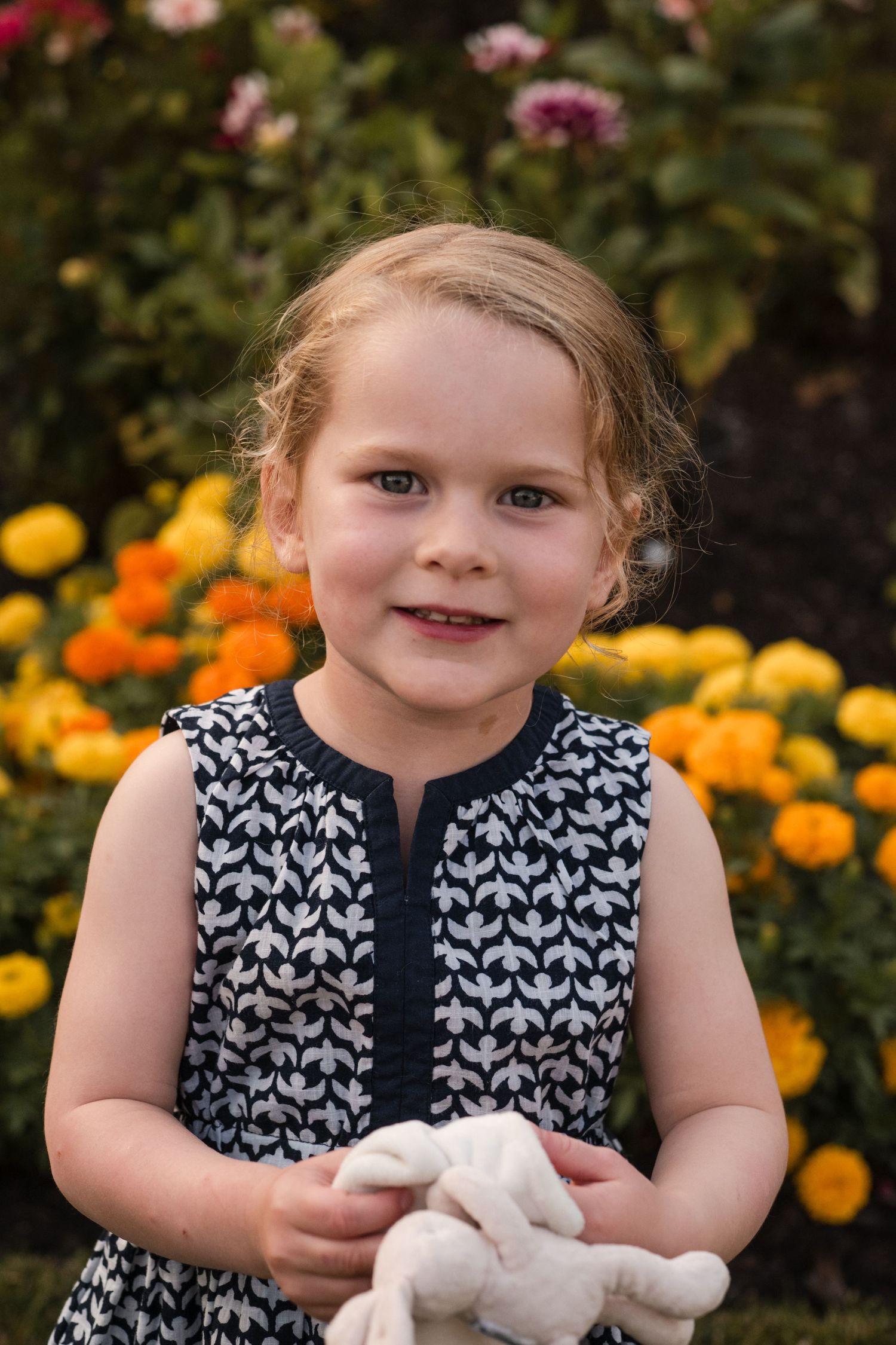 A child in a black and white patterned dress poses with a stuffed animal in a garden with orange marigolds.