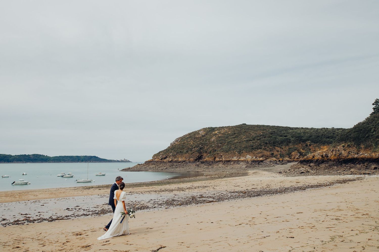 Bride in white wedding dress walking alone on sandy beach with rocky coastline and calm sea.
