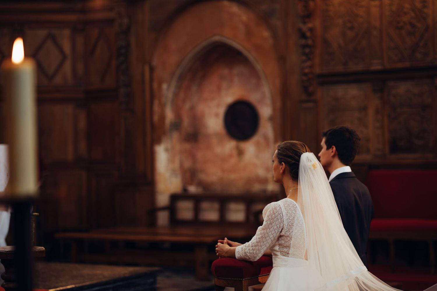 Bride and groom sitting at church altar with ornate stone architecture and glowing candle nearby.