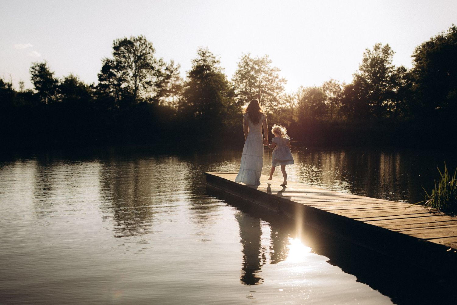 Silhouettes stand on wooden dock at sunset with sun reflecting off calm lake waters and trees in background.