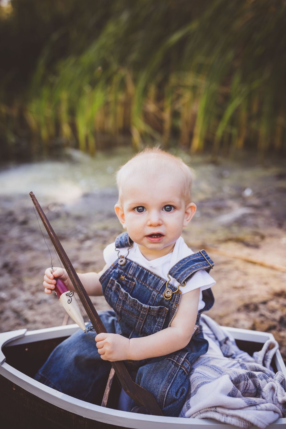 A baby in denim overalls sits in a rustic wooden wagon on a sunny day near tall grass.