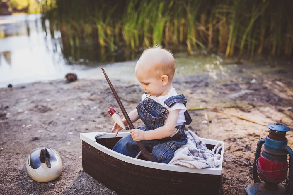 A baby plays in the sand at sunset while wearing blue denim overalls and a striped shirt.