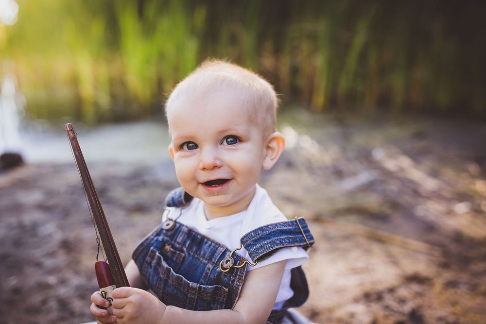 Small child in denim overalls holding a stick while playing outdoors near tall grass and water.