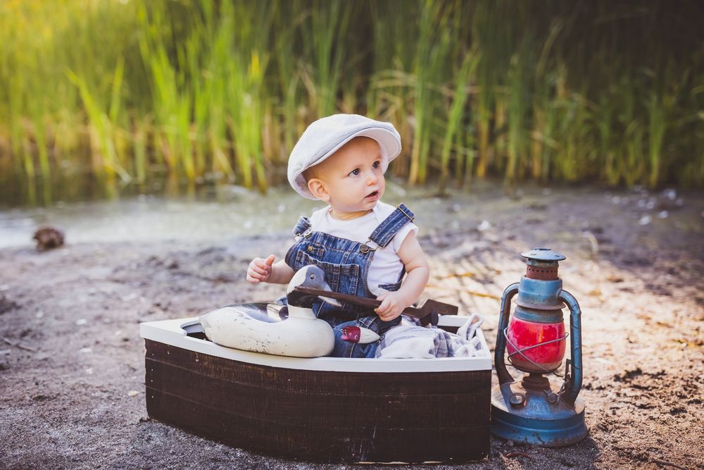 Baby in overalls sits on blanket with vintage lantern during outdoor photoshoot.