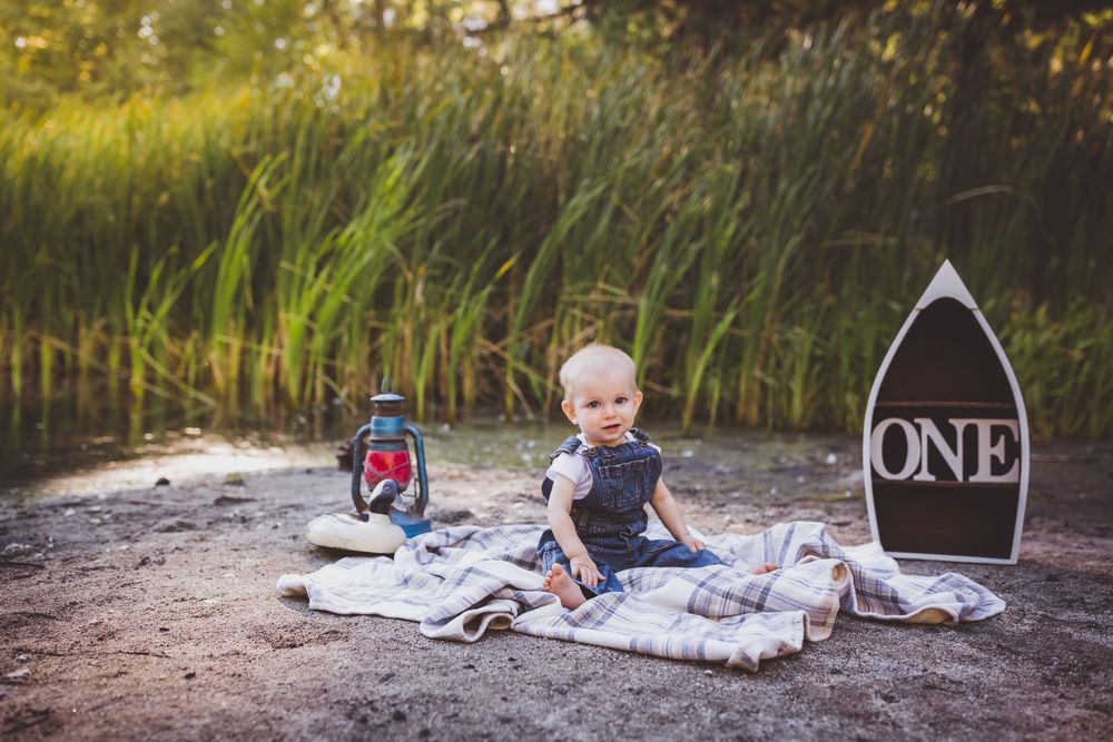 Baby on blanket with lantern and 'ONE' sign for first birthday photoshoot by water.