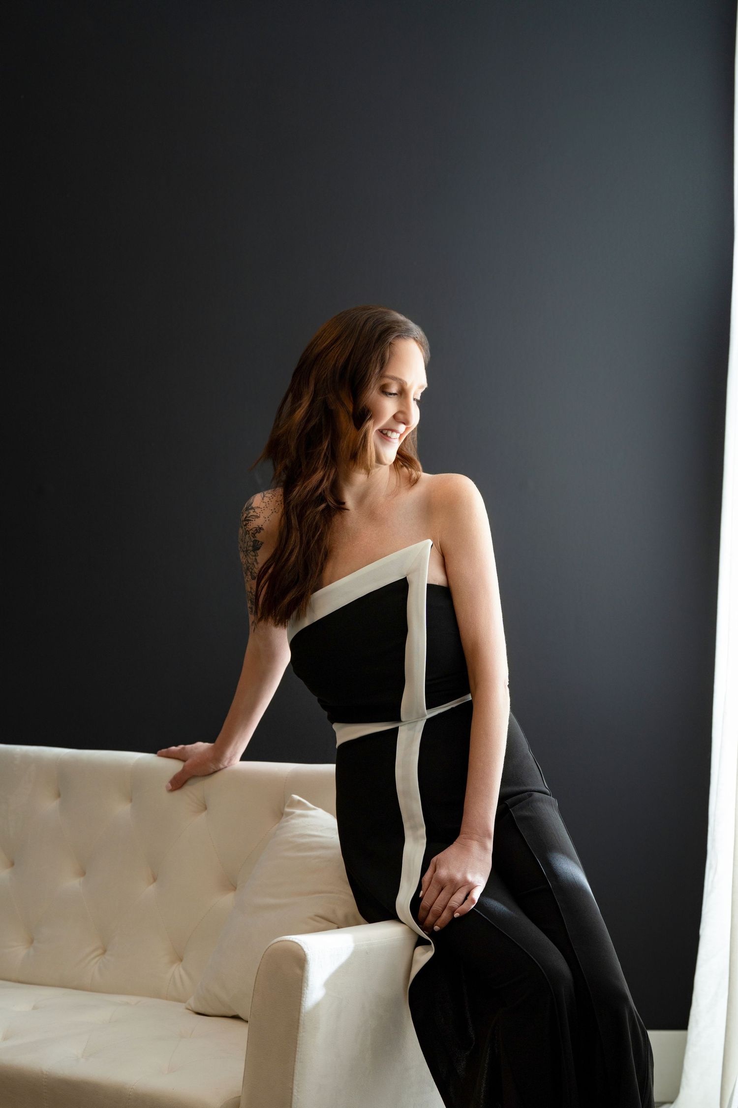 A woman in a black dress poses elegantly on a cream-colored tufted sofa against a dark gray backdrop.
