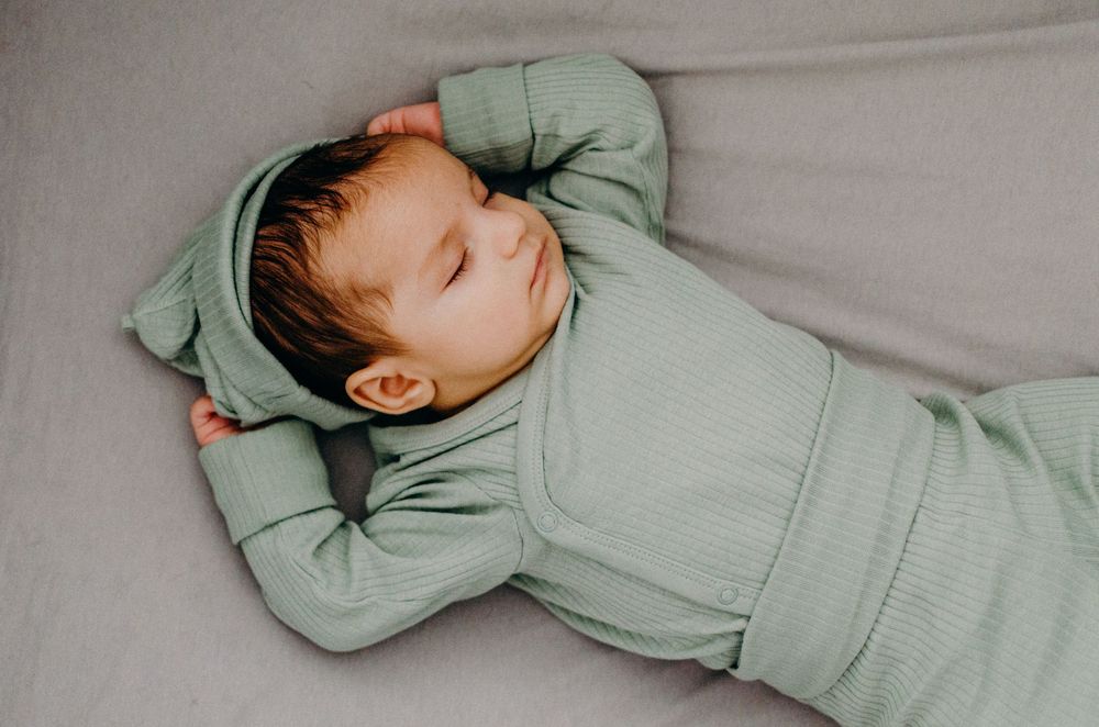 A sleeping baby wearing a cozy mint green outfit lies peacefully on light gray bedding.