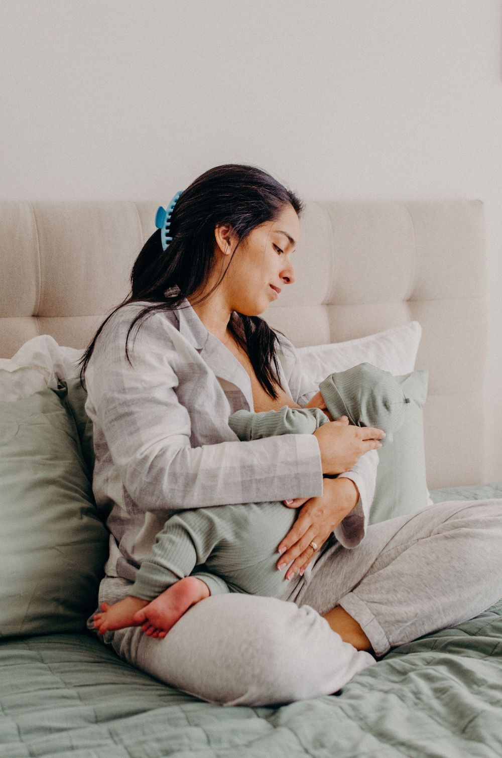 Woman in gray loungewear cuddles with infant on comfortable bed with neutral bedding.