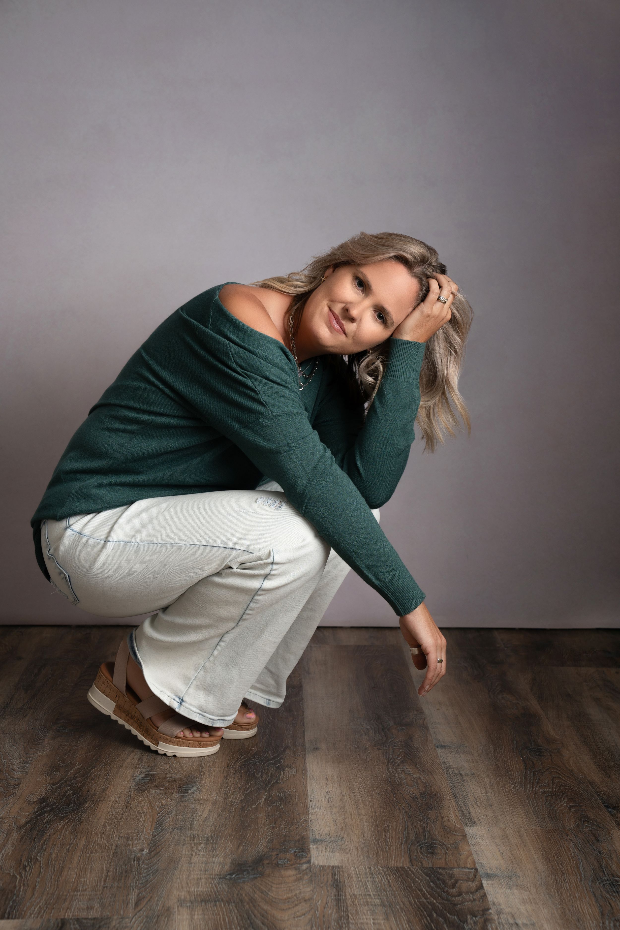 Person in green sweater and white pants crouches in dynamic poses against gray backdrop on wooden floor.