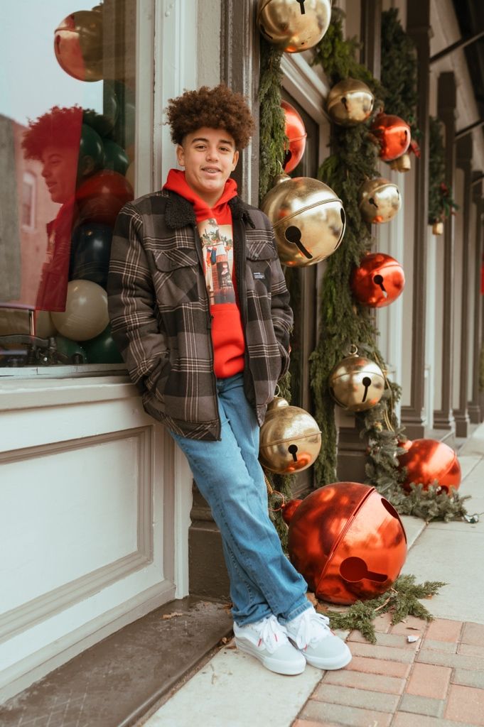 Person in plaid jacket and jeans poses next to holiday decorations and greenery outside storefront window.