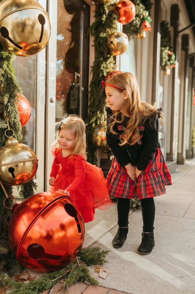 Children in festive outfits play near holiday decorations and large red ornaments on sidewalk.
