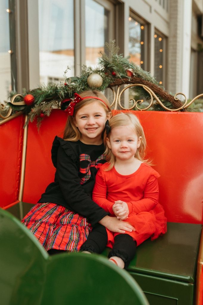 Two children in festive holiday outfits sitting on a red bench decorated with Christmas greenery and ornaments.