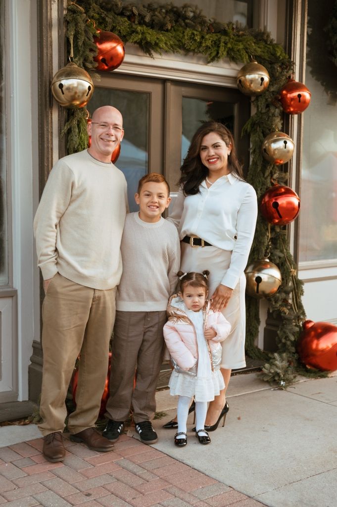 Family wearing coordinated white and beige outfits poses for holiday photos in front of decorated storefront.