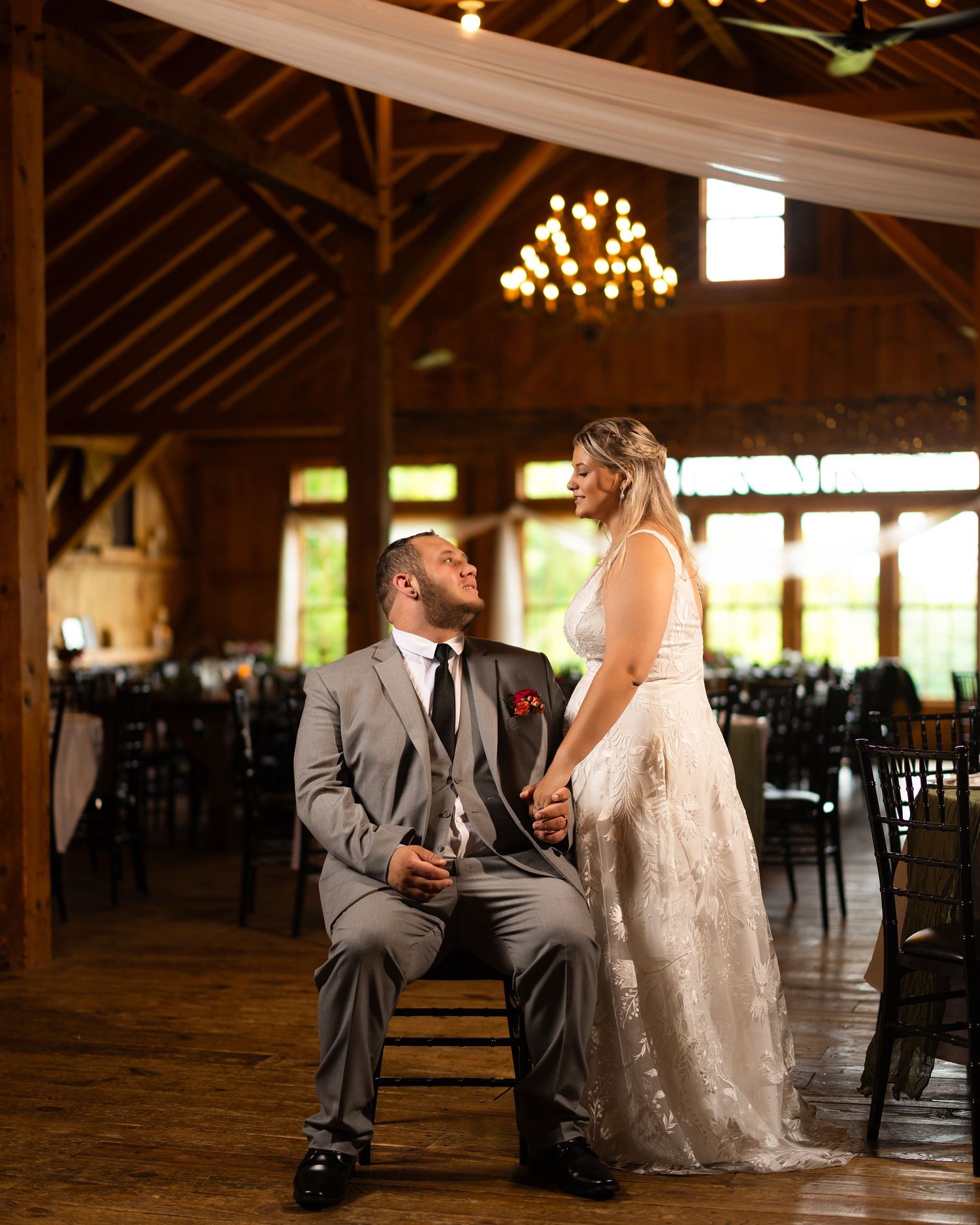 Bride and groom share a romantic moment in a rustic barn wedding venue with wood beams and chandelier lighting.