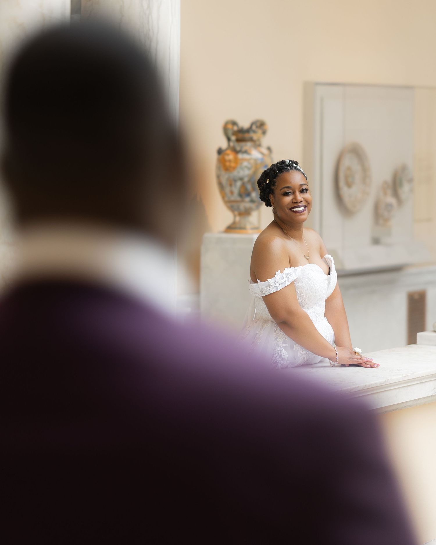 Smiling bride in white off-shoulder gown looks toward groom during wedding ceremony in elegant venue.