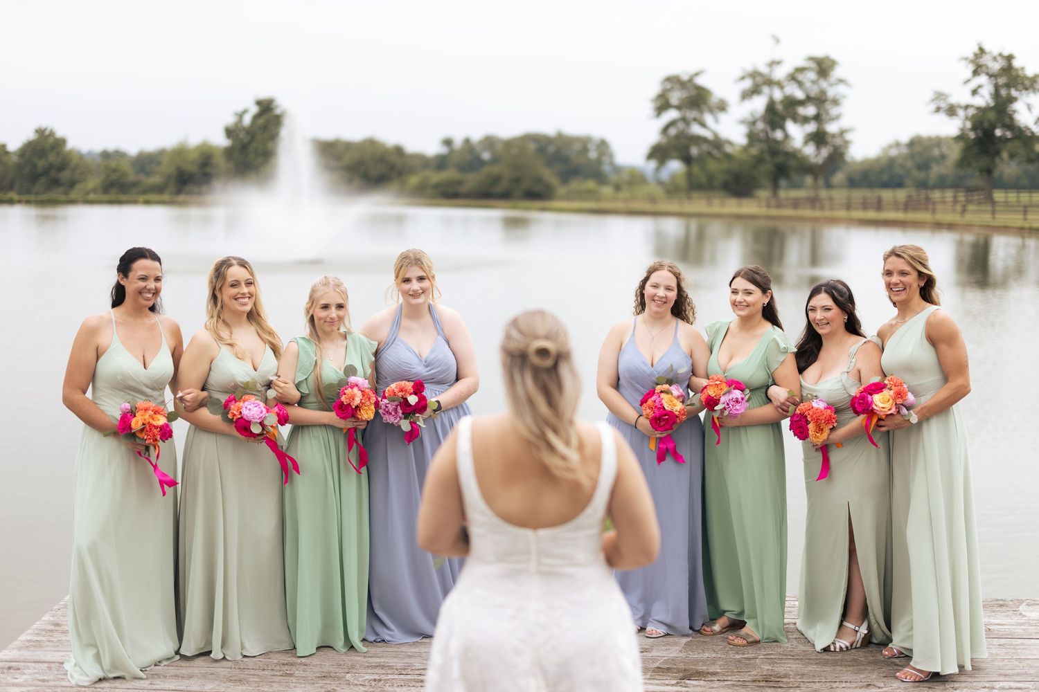 Bride faces her bridesmaids in sage and lavender gowns holding vibrant bouquets on a dock by a serene pond.