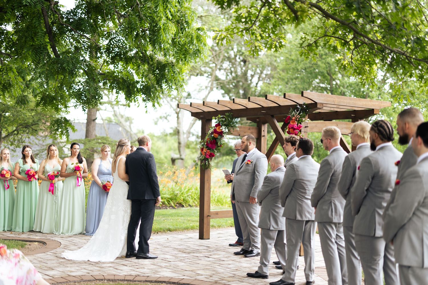 Outdoor wedding ceremony under trees with bride, groom, officiant at wooden pergola altar and wedding party.